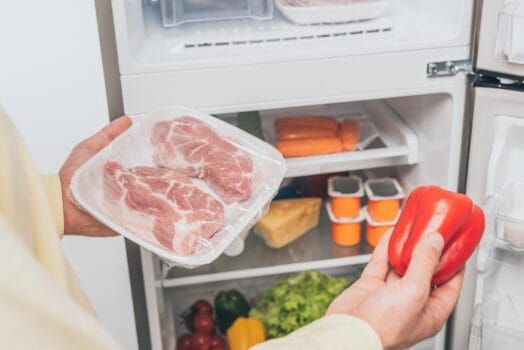 男性手拿冷凍肉與甜椒站在裝滿食物的冰箱前 / Man holding frozen meat and bell pepper near an open well-stocked fridge