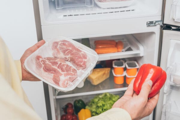 男性手拿冷凍肉與甜椒站在裝滿食物的冰箱前 / Man holding frozen meat and bell pepper near an open well-stocked fridge